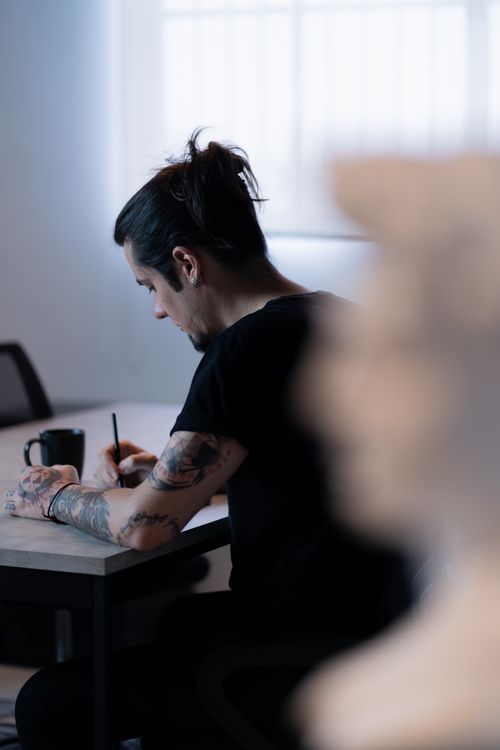 Free A thoughtful tattooed man writing at his desk in a modern office setting. Stock Photo