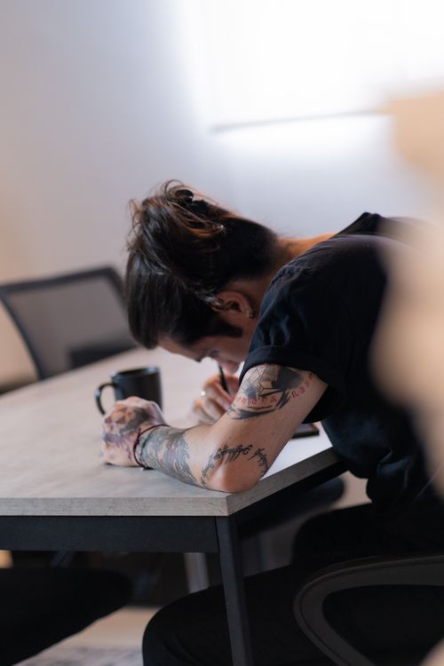 Free A tattooed individual rests their head on a desk beside a mug in a dimly lit room. Stock Photo