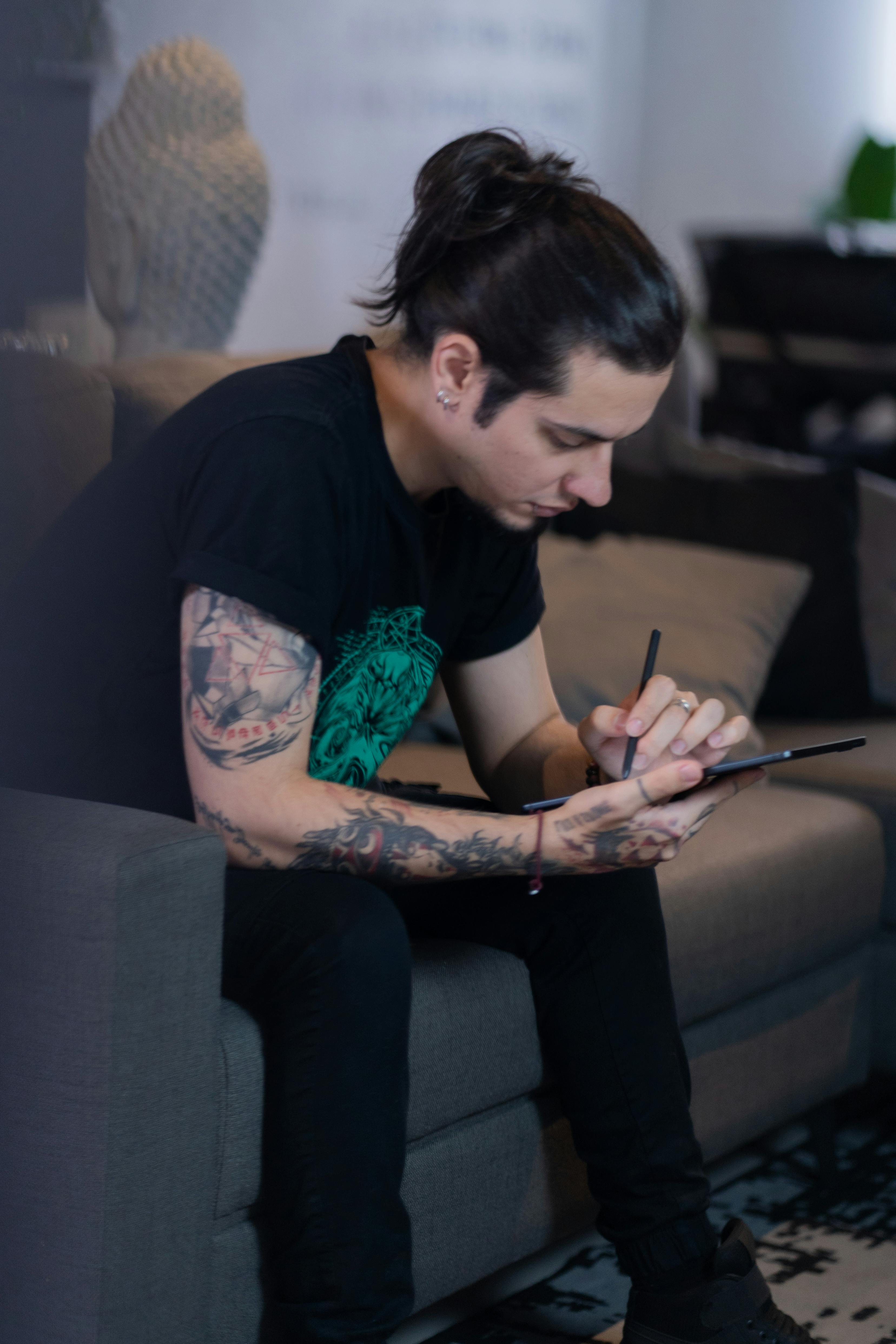 免費 紋身男子坐在室內的沙發上，在平板電腦上進行數位繪畫。 圖庫相片