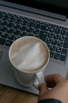 A hand holding a cappuccino cup on a laptop keyboard, combining work and coffee break.