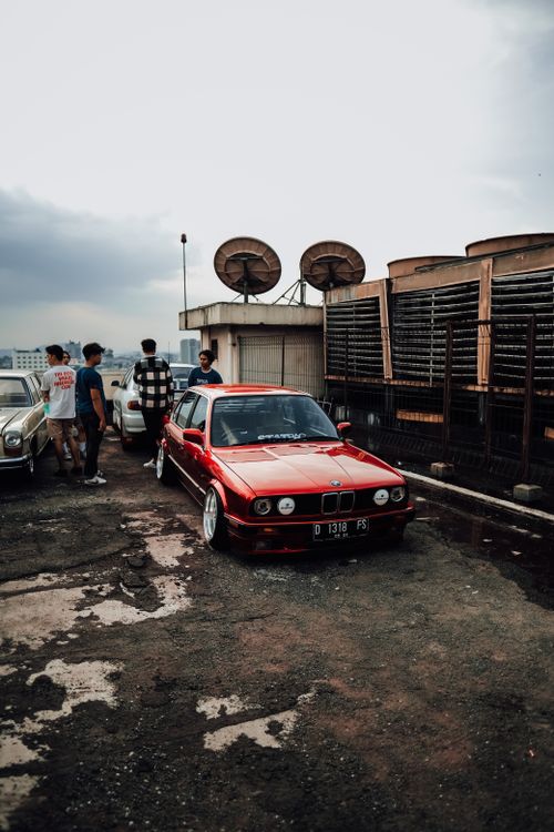 Free A red vintage car parked on a rooftop with people socializing and cityscape in the background. Stock Photo