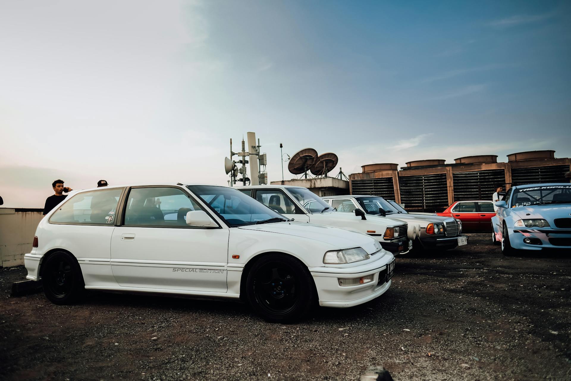 A lineup of classic cars parked outdoors with people nearby under a clear sky.