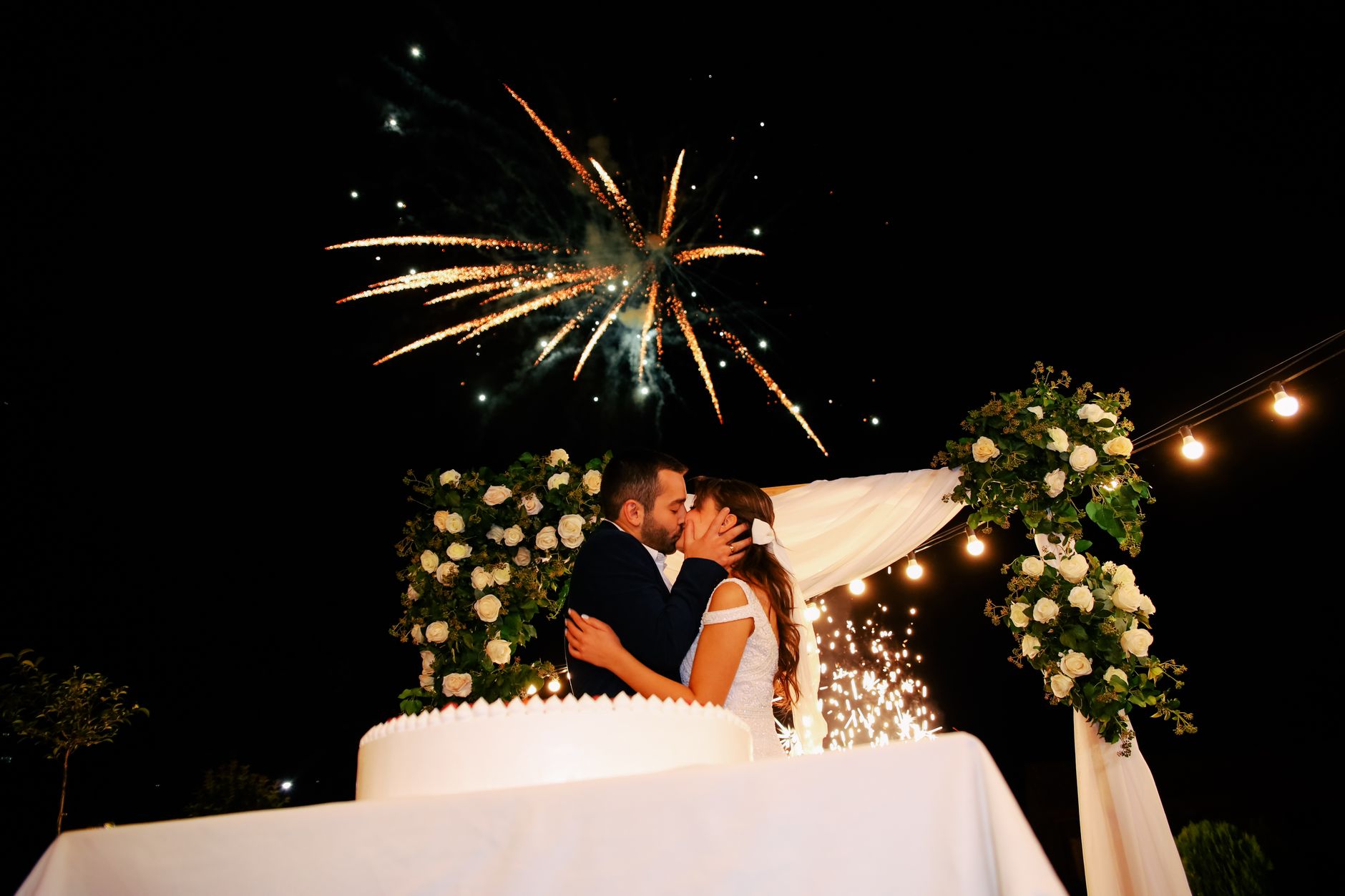 Romantic outdoor wedding scene with couple kissing under a beautiful fireworks display.