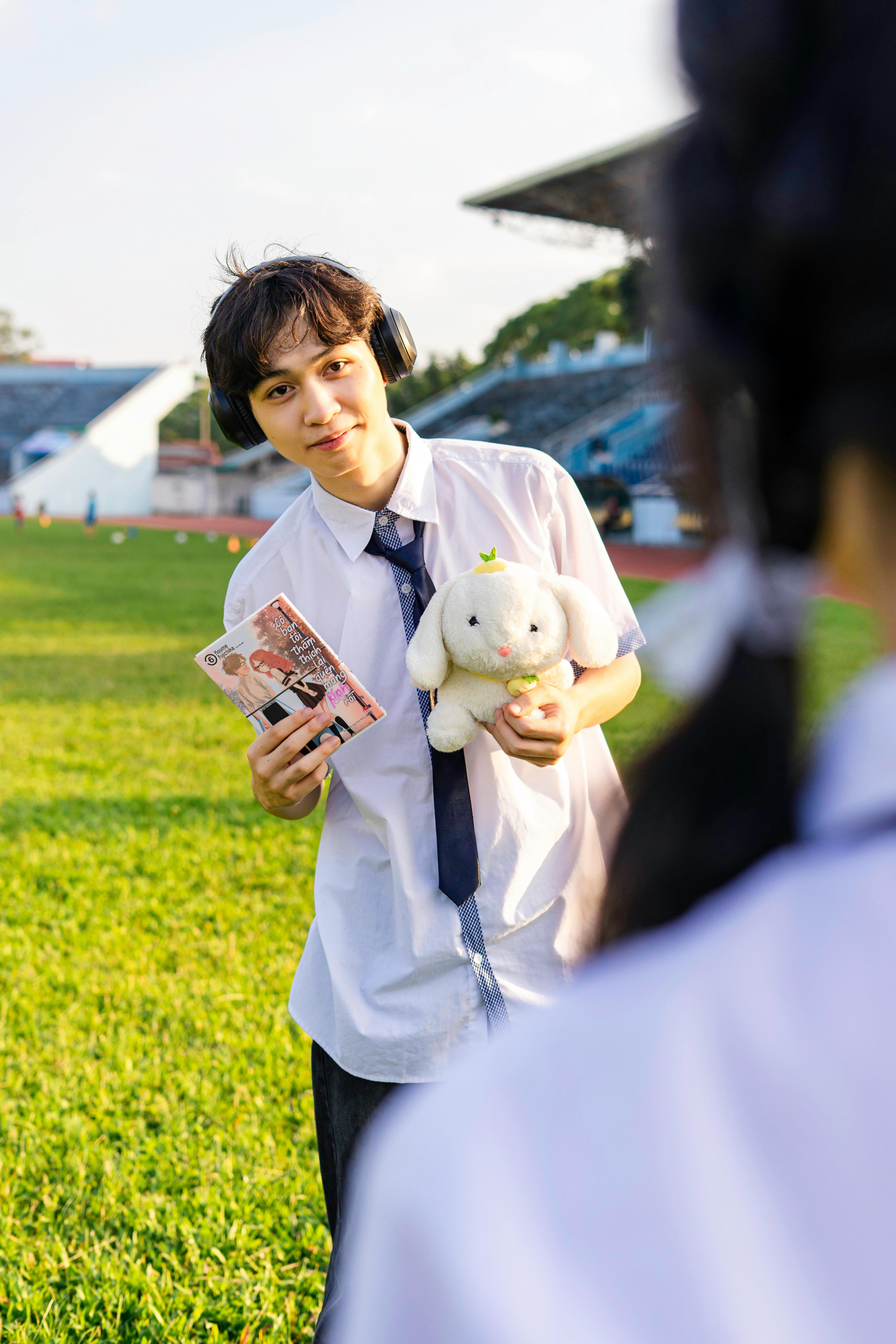 Teen boy holding a stuffed animal and headphones outdoors in a field, smiling.