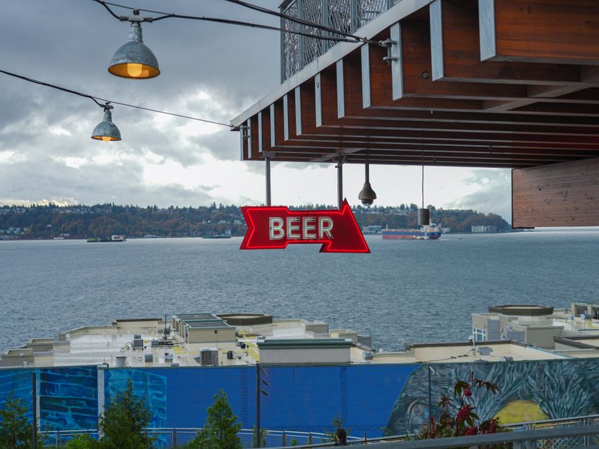 Neon beer sign hanging over waterfront with industrial and scenic views.