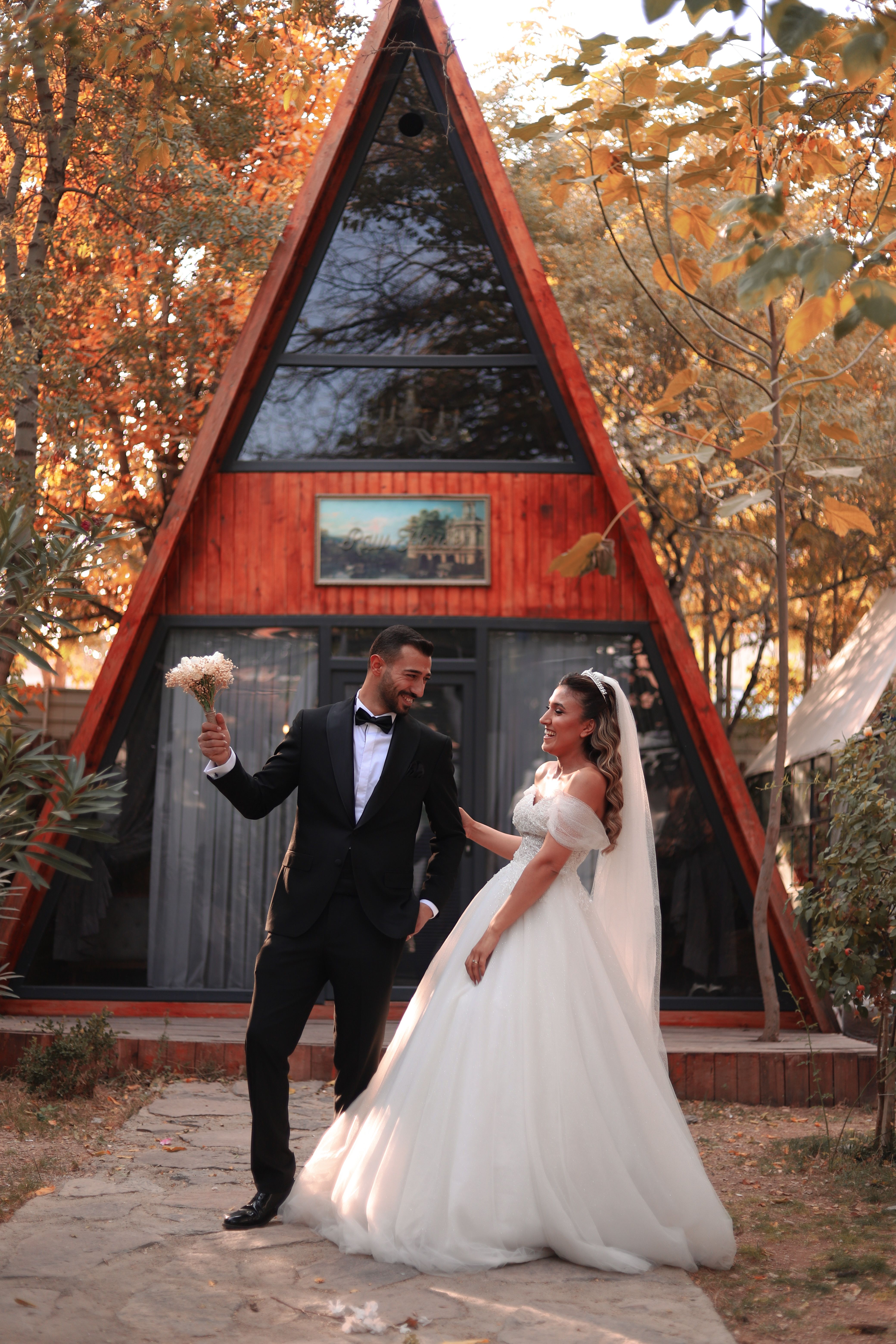 Bride and groom pose joyfully in front of a charming A-frame cabin under autumn leaves.