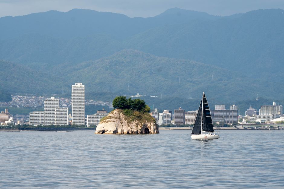 Sailboat cruising past a small island with a cityscape and mountain backdrop.