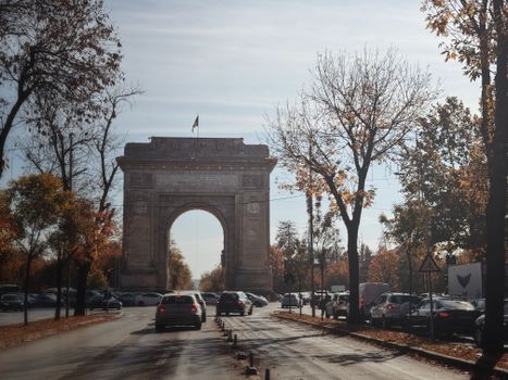 Triumphal arch in Bucharest surrounded by autumn trees and traffic.