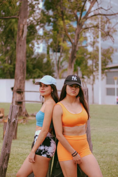 Free Two young women in activewear leaning against a fence outdoors amid greenery and trees. Stock Photo