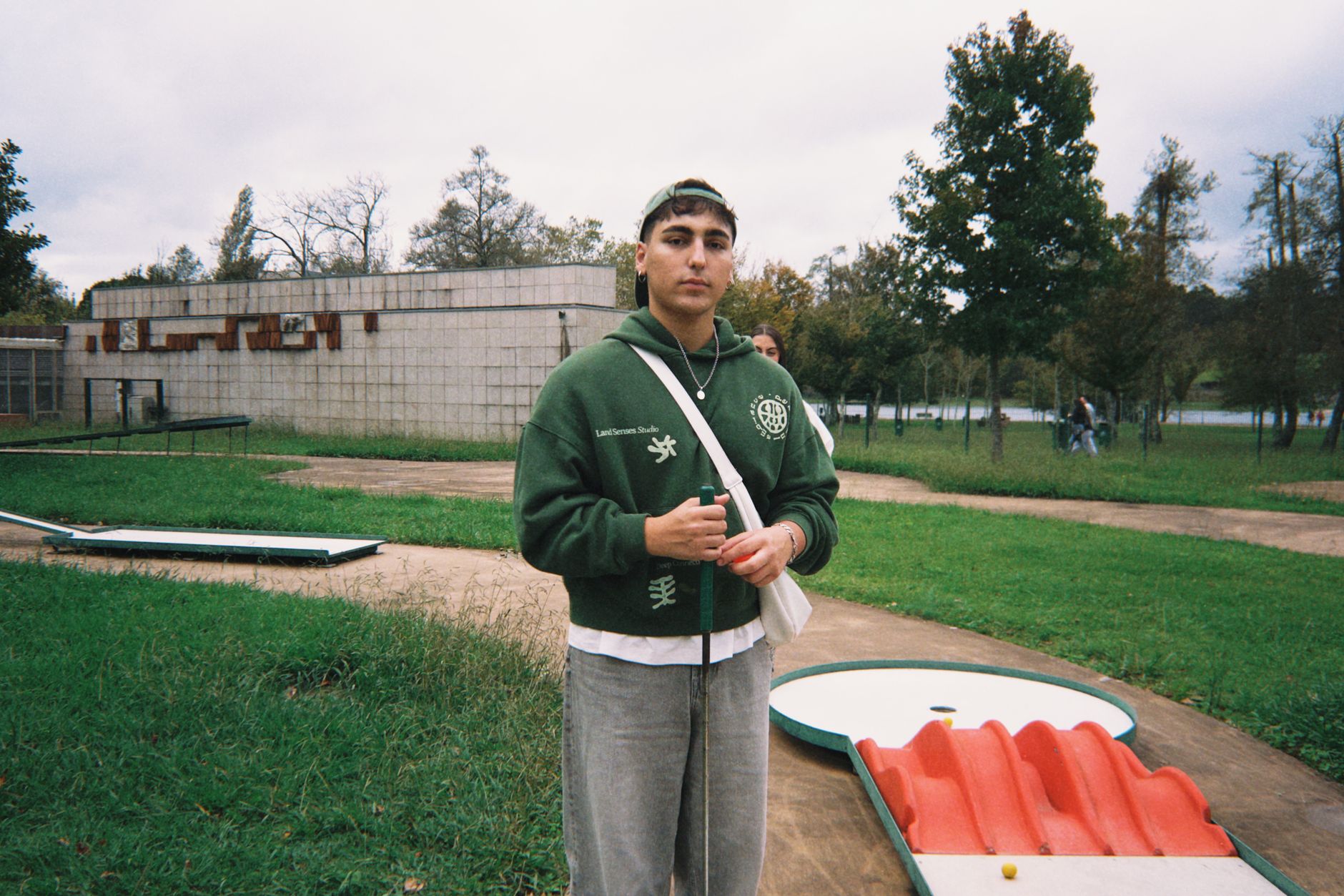 A young man stands at a mini-golf course outdoors in casual attire.
