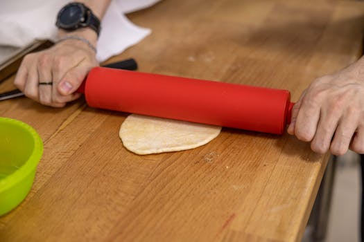 Close-up of a person rolling dough with a red rolling pin on a wooden surface.