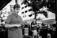 在充满活力的城市街道上，一位年轻的街头音乐家弹奏着四弦琴，街道两旁摆满了户外市场摊位