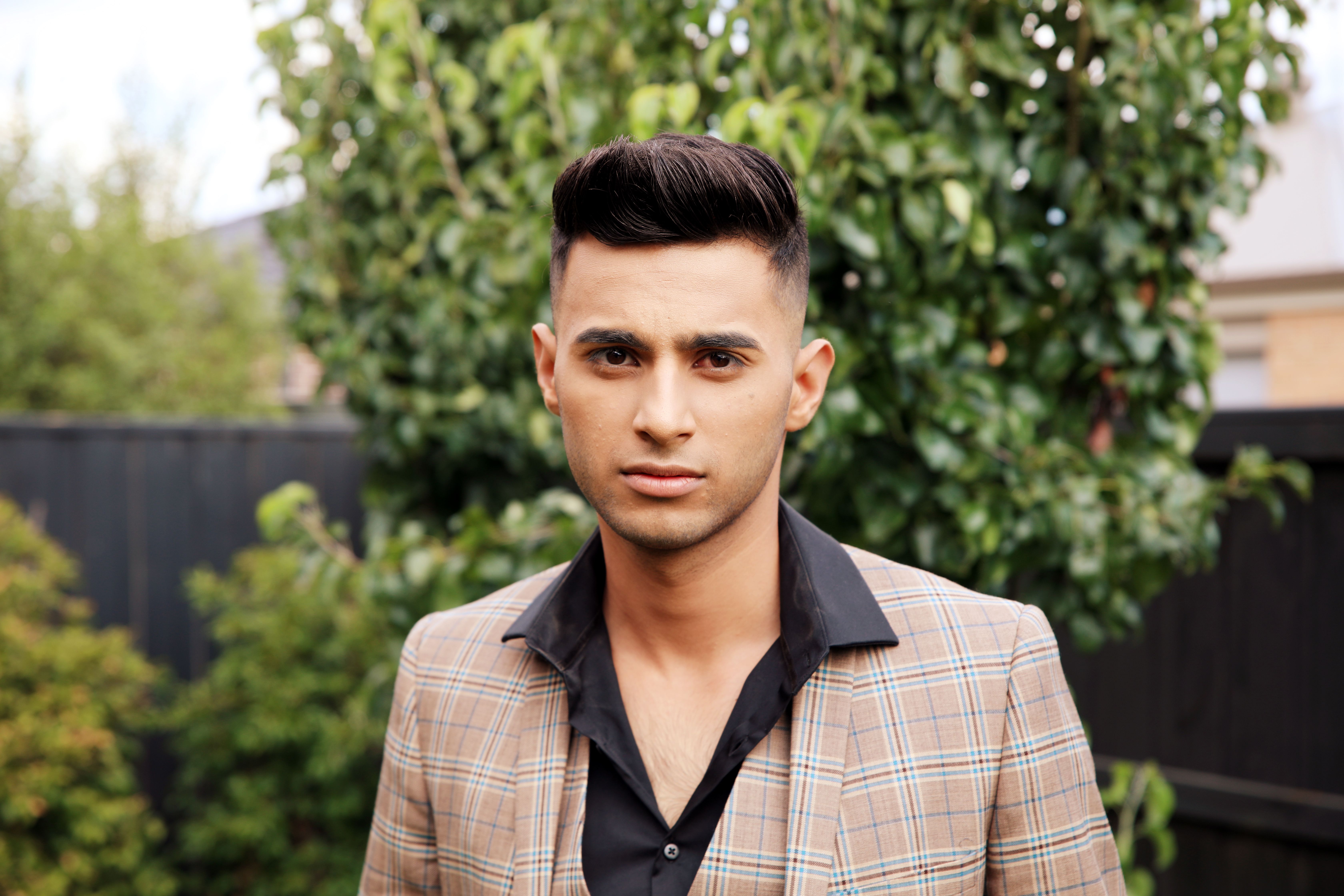 Confident young man in a plaid jacket poses outdoors against a leafy background.