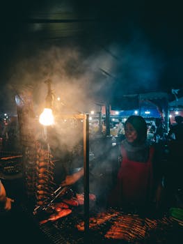 A woman in a hijab grills traditional food at a bustling night market, surrounded by smoke.