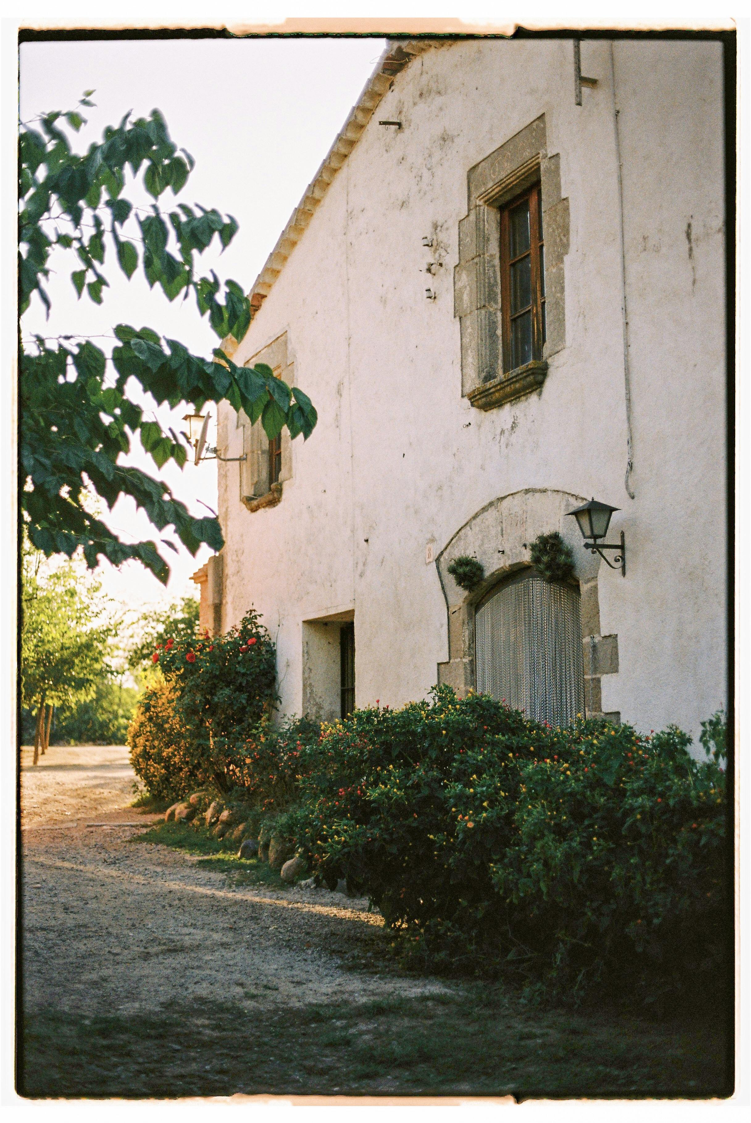 A rustic house with wooden shutters surrounded by a lush garden in warm sunlight.