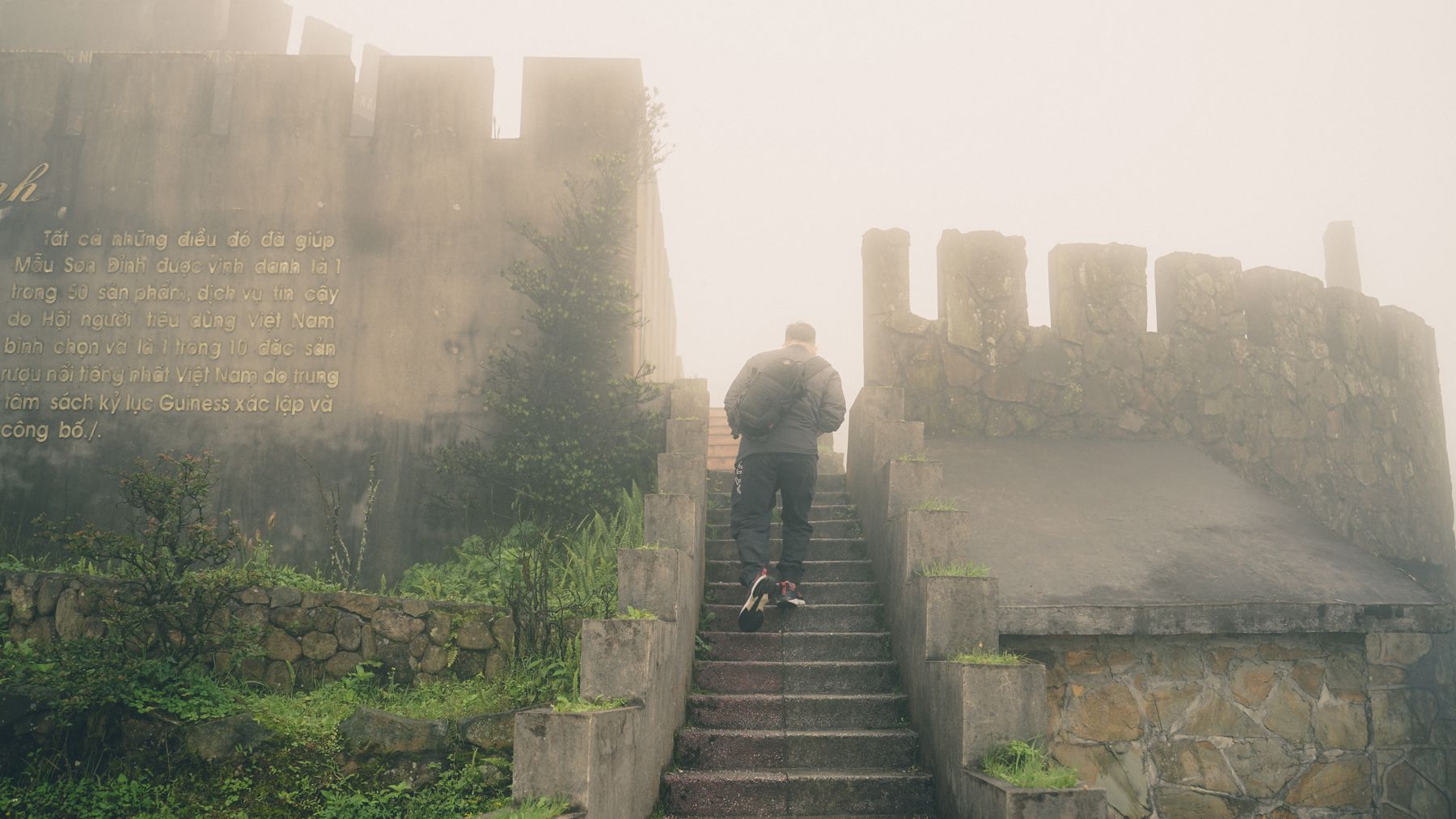 A man ascends a misty stone staircase surrounded by fog and ancient walls, capturing a mysterious atmosphere.