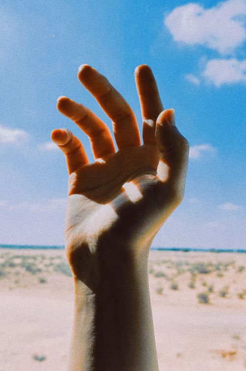Free A hand reaching out towards a sunny blue sky with a desert landscape in the background. Stock Photo