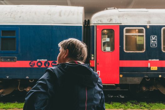 Man waiting by a red and blue train at an outdoor station, viewed from behind.