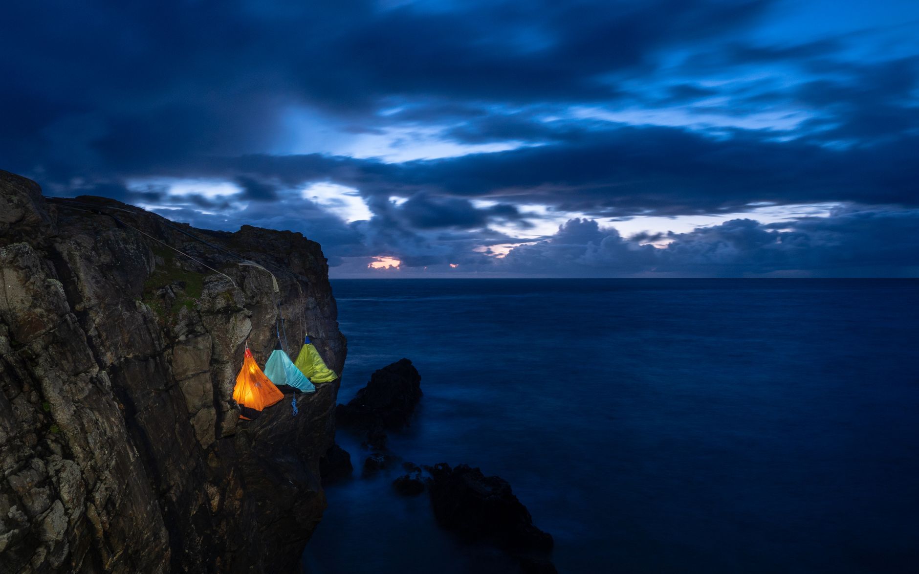 Colorful tents hang on a cliffside against a stunning twilight ocean view, creating an adventurous and serene atmosphere.