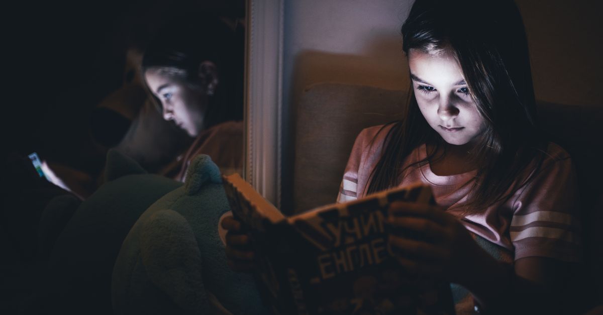 A young girl reading a book in a dimly lit room, reflecting in a mirror.