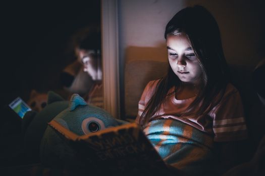 A young girl reads a book at night with subtle lighting, reflecting a calm and cozy atmosphere.