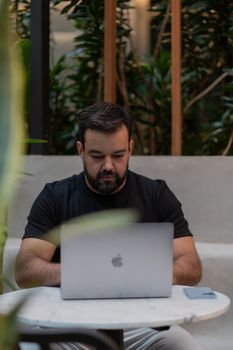 Adult man focused on his laptop while seated in a modern cafe with plants.