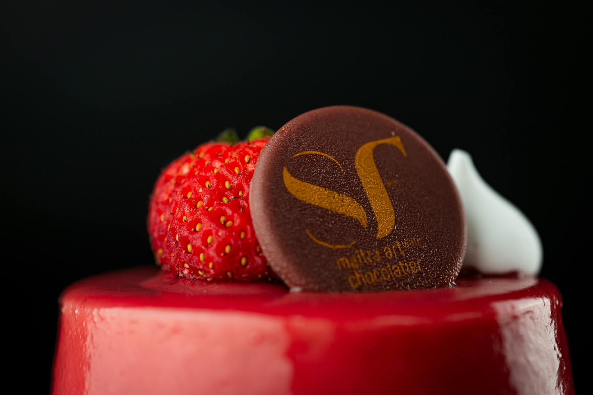 Closeup of a red glazed cake topped with fresh strawberry and branded chocolate disc.