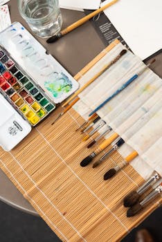 A detailed view of an artist's workspace featuring watercolor paints and brushes on a bamboo mat.