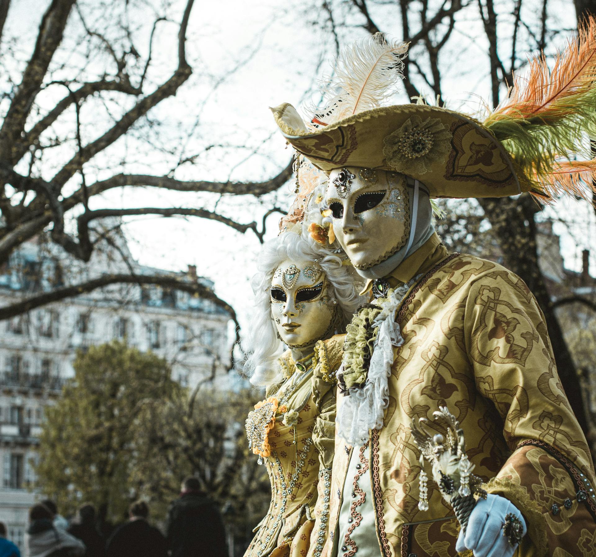 Two costumed individuals in traditional Venetian carnival attire with ornate masks and hats, set in an outdoor urban environment.