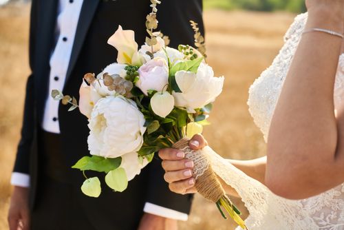Free Beautiful floral arrangement held by bride in lace gown, alongside groom in suit, outdoors on sunny day. Stock Photo