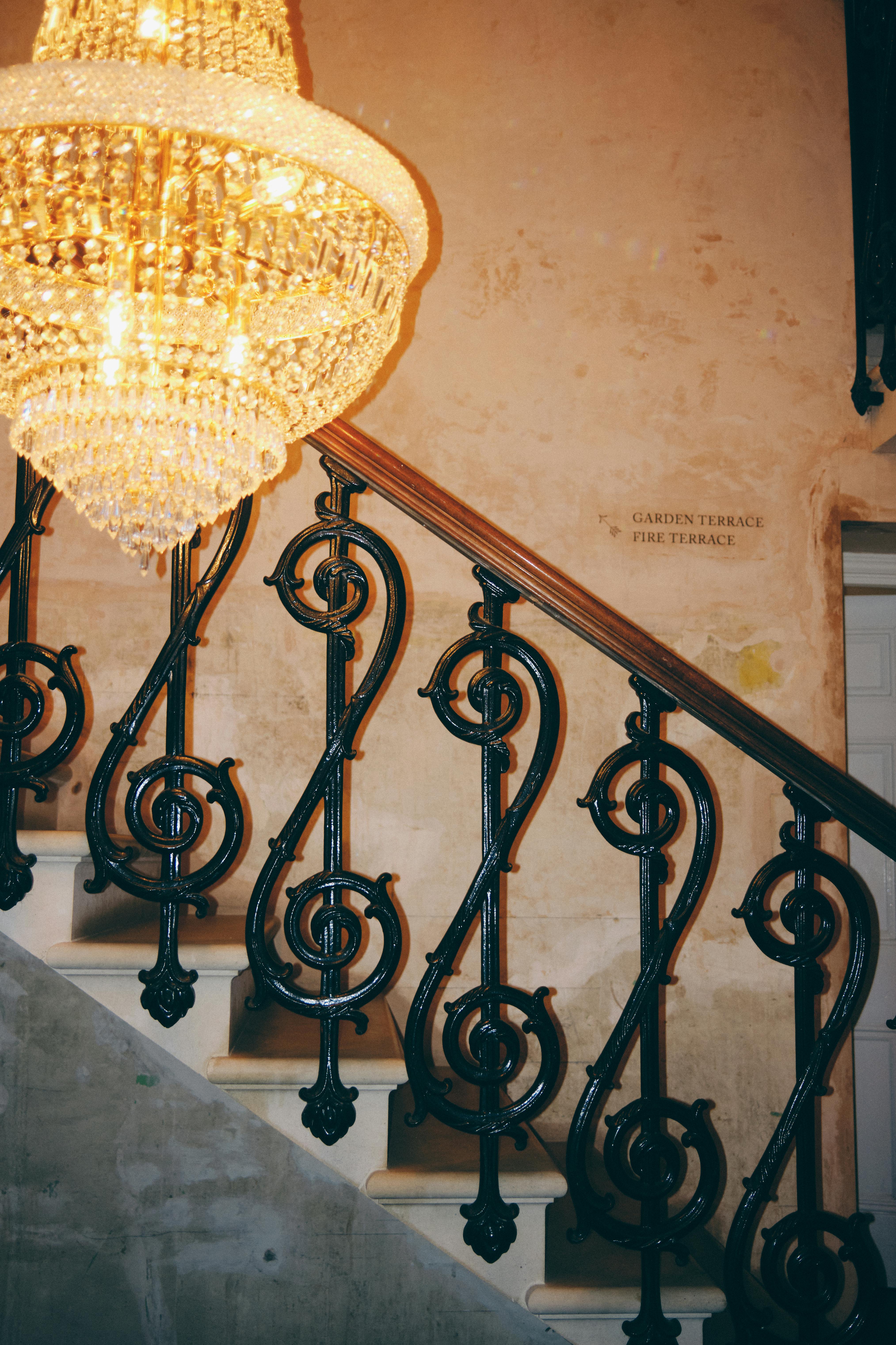 Luxurious chandelier illuminating vintage staircase with ornate iron railing.