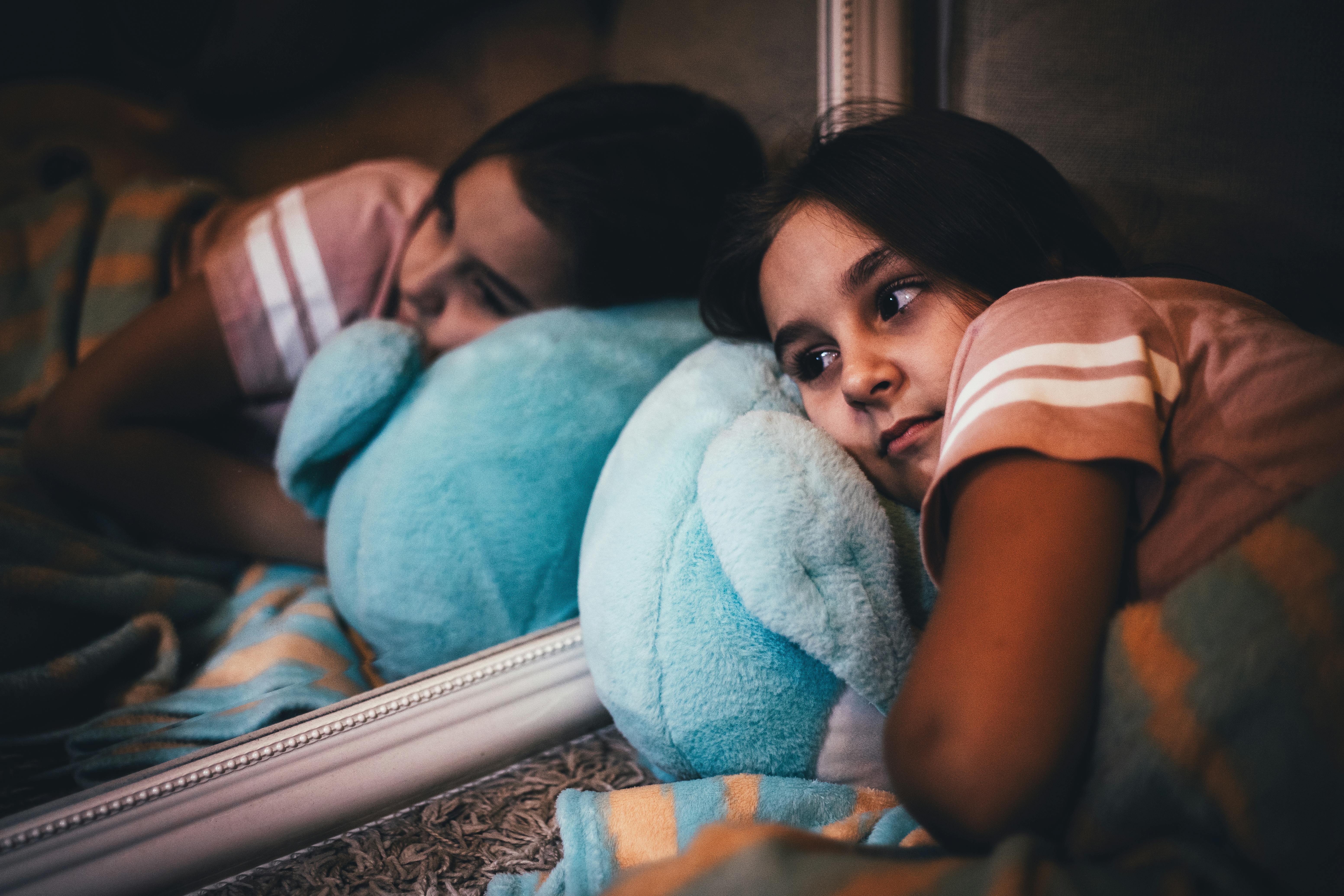 Young girl lying on a bed with a plush toy, reflecting in a mirror, deep in thought.