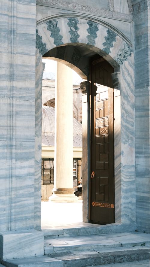 Free Intricate marble archway leading to a sunlit courtyard through an open wooden door. Stock Photo
