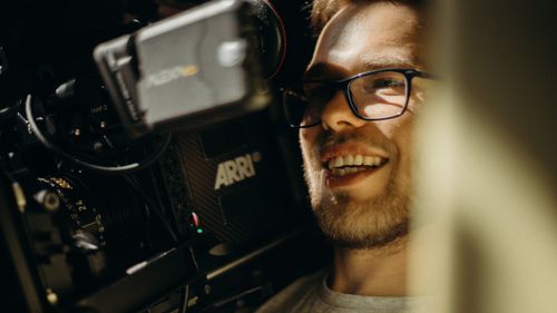 Free A smiling filmmaker with eyeglasses operates an ARRI camera, capturing a lively moment on set. Stock Photo
