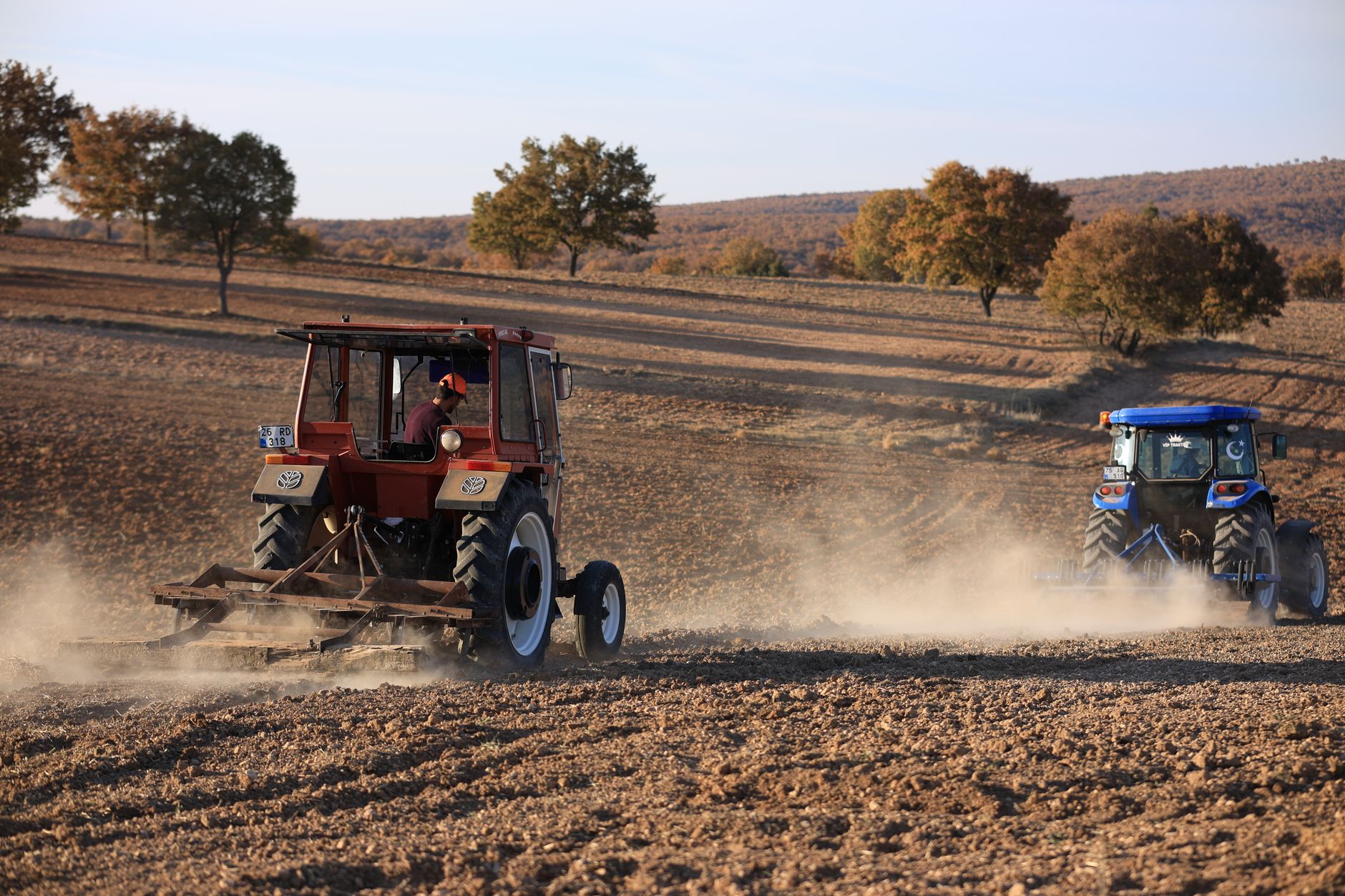 Two tractors plowing a dusty field in a rural autumn landscape.
