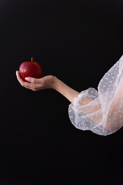 A delicate hand in a sheer sleeve holds a red apple against a black background.