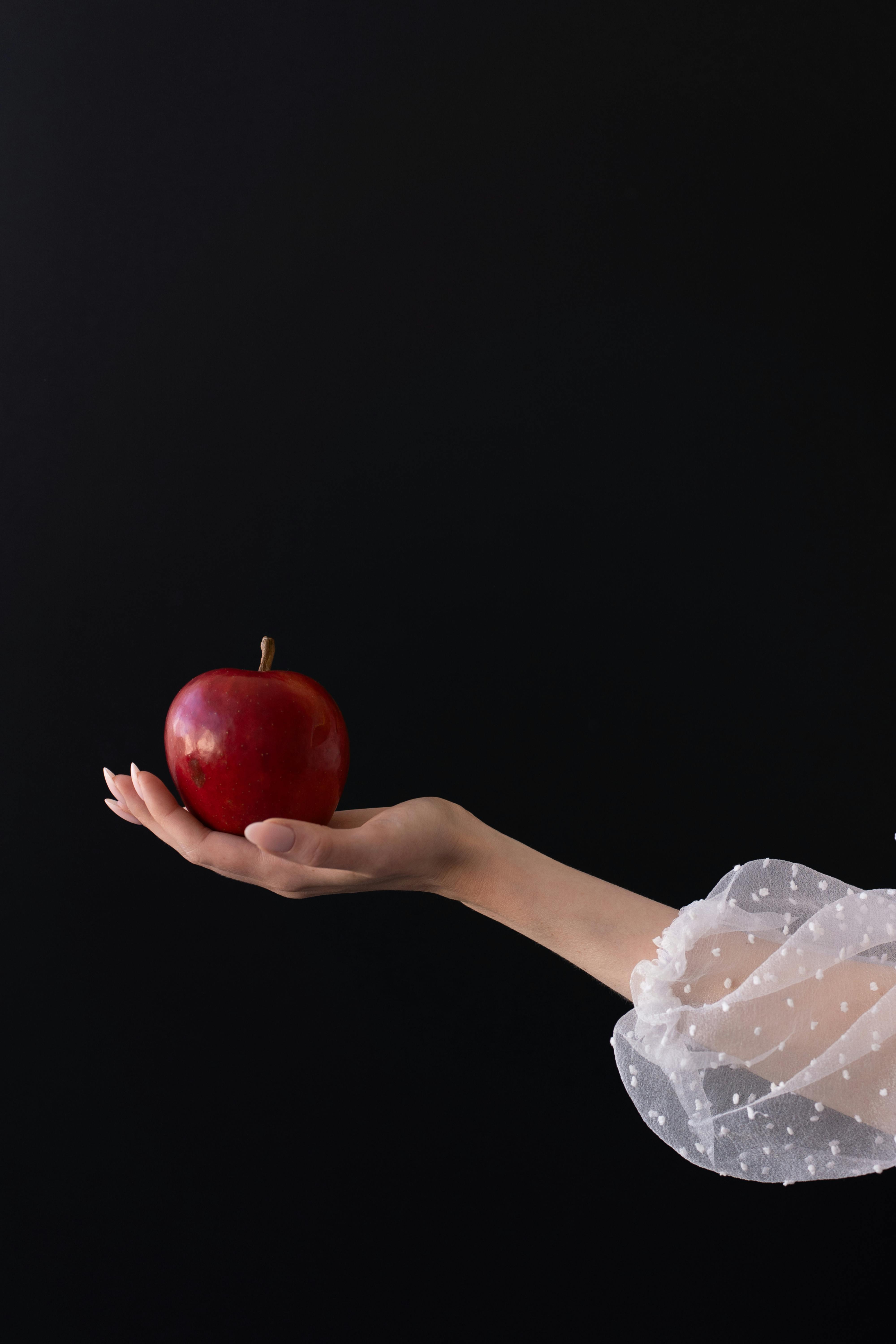 A delicate hand presenting a red apple, symbolizing themes of temptation and health.