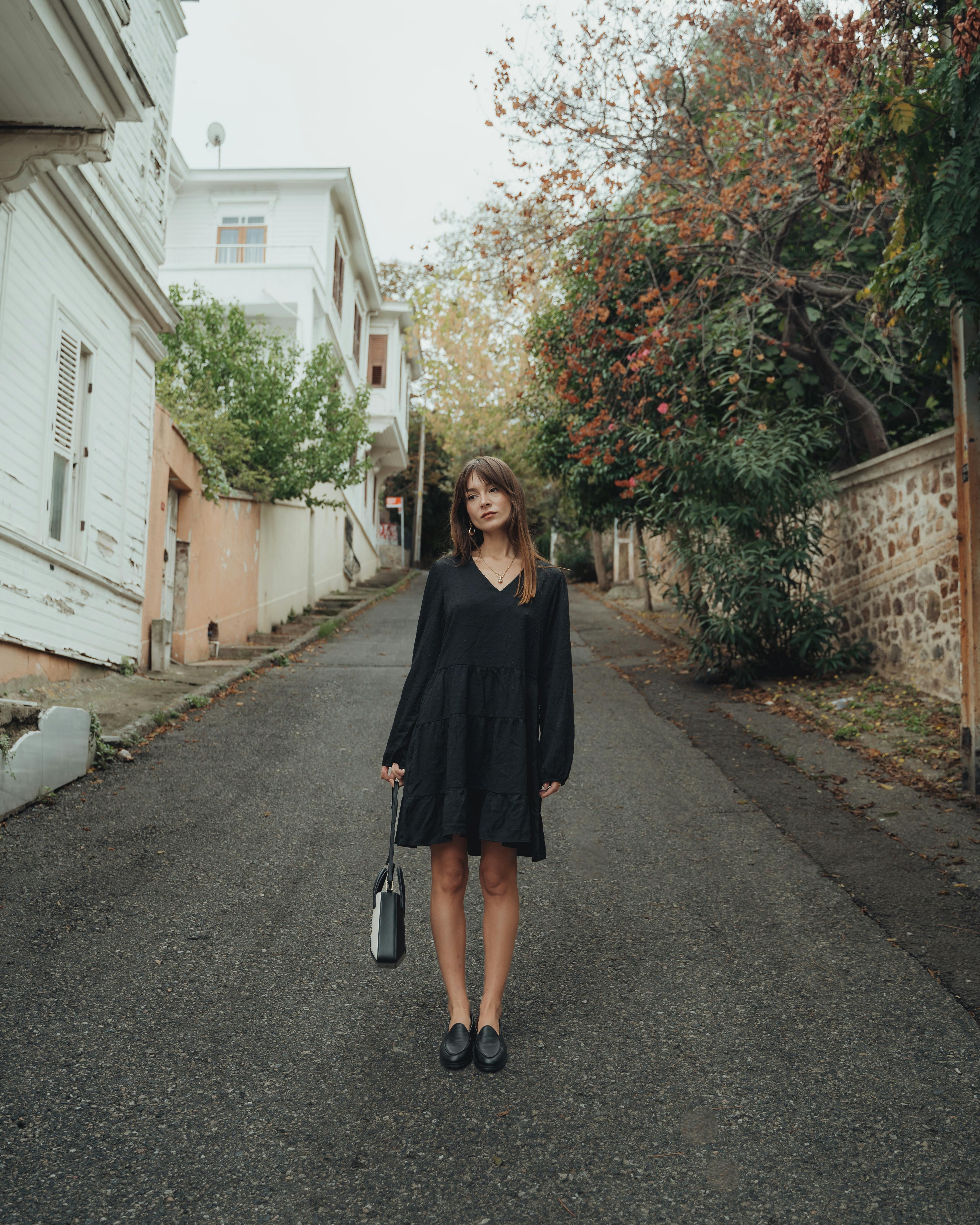 Young woman in a black dress stands alone on a quiet street surrounded by charming buildings.