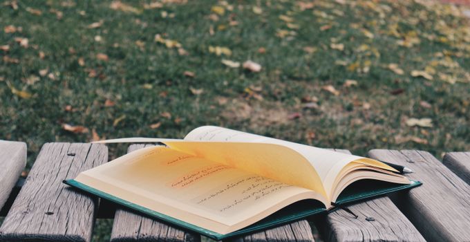 An open book with text on a bench in a park setting during fall.