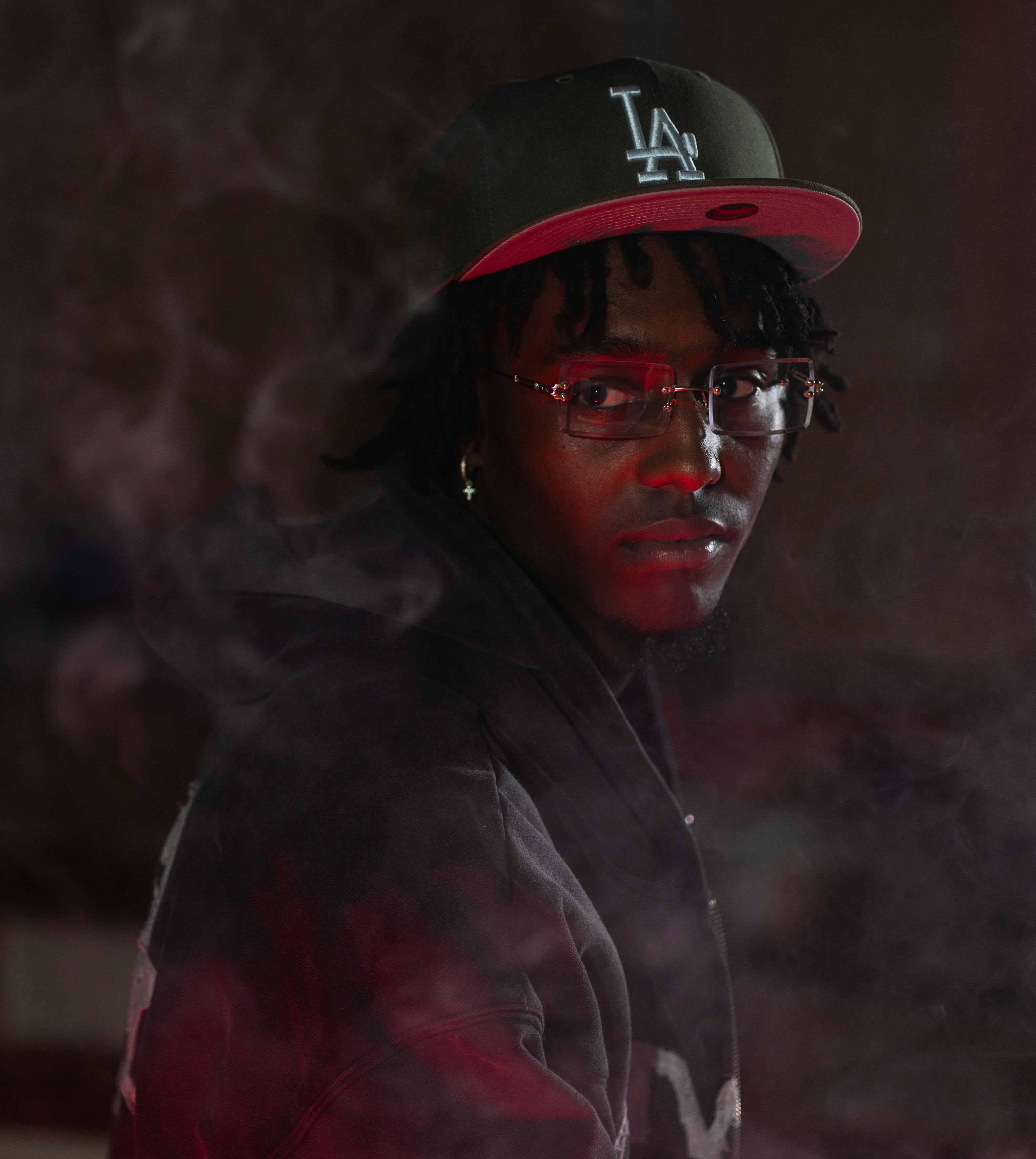 A moody portrait of a young man in glasses and a cap amidst smoke, shot indoors.