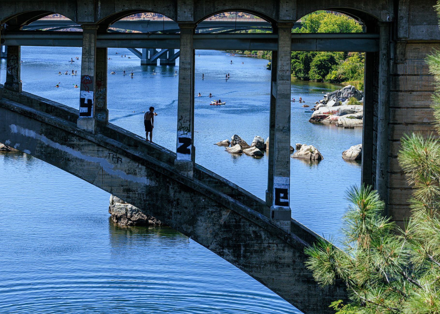 A person stands silhouetted on the Rainbow Bridge in Folsom, overlooking a serene lake.