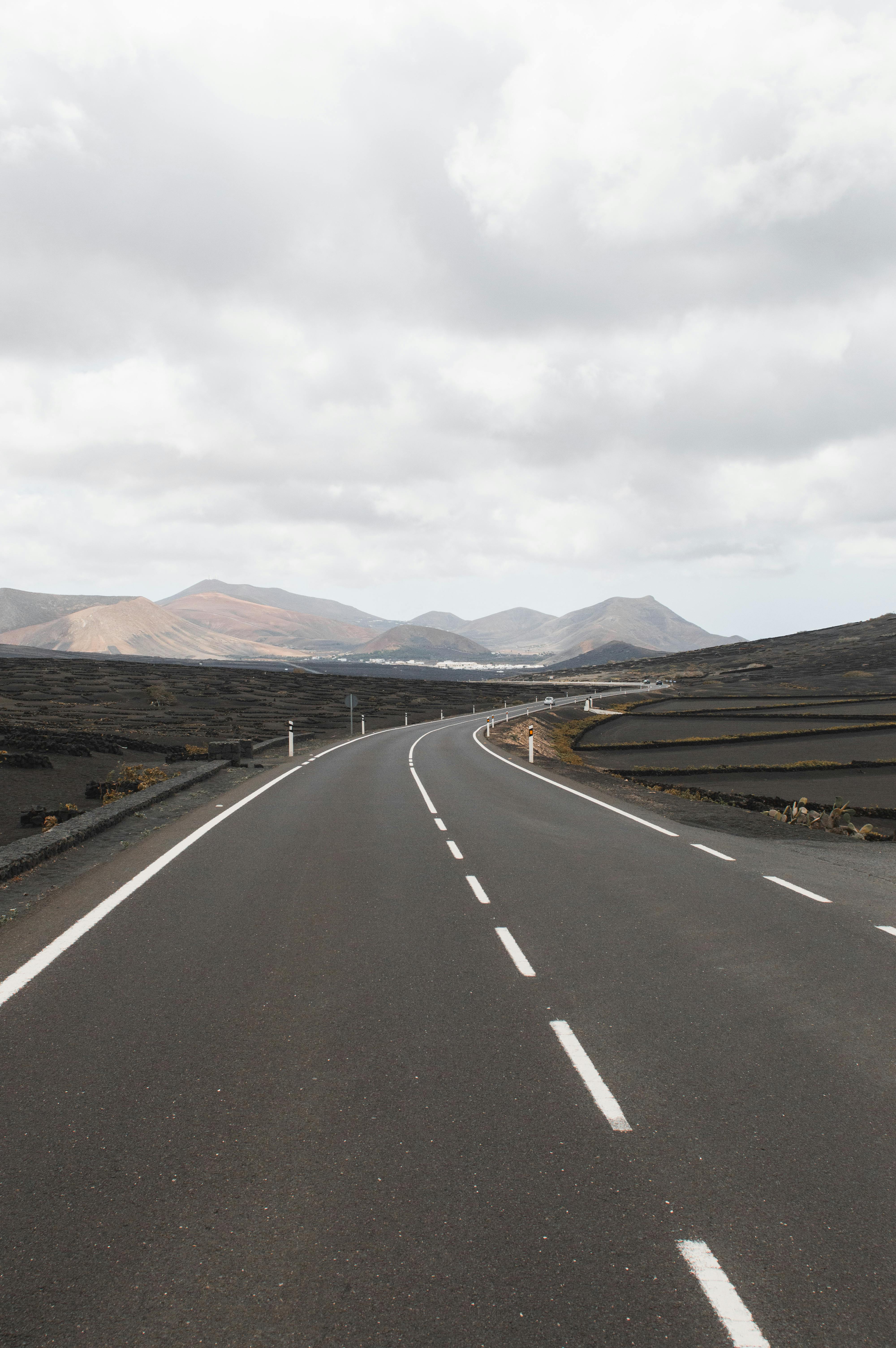 Curved road through the volcanic landscape of Lanzarote, Spain, with mountains in the distance under an overcast sky.
