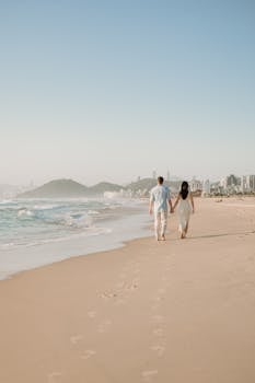 A couple walking hand-in-hand on a sunny beach, leaving footprints in the sand.