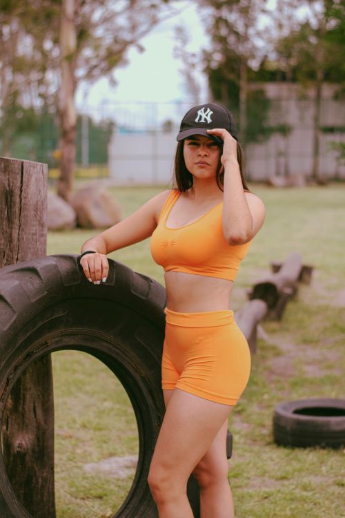 Free Confident woman in orange sportswear posing by tire in an outdoor setting. Stock Photo