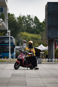 A delivery rider in uniform takes a break on a red scooter in a city area.