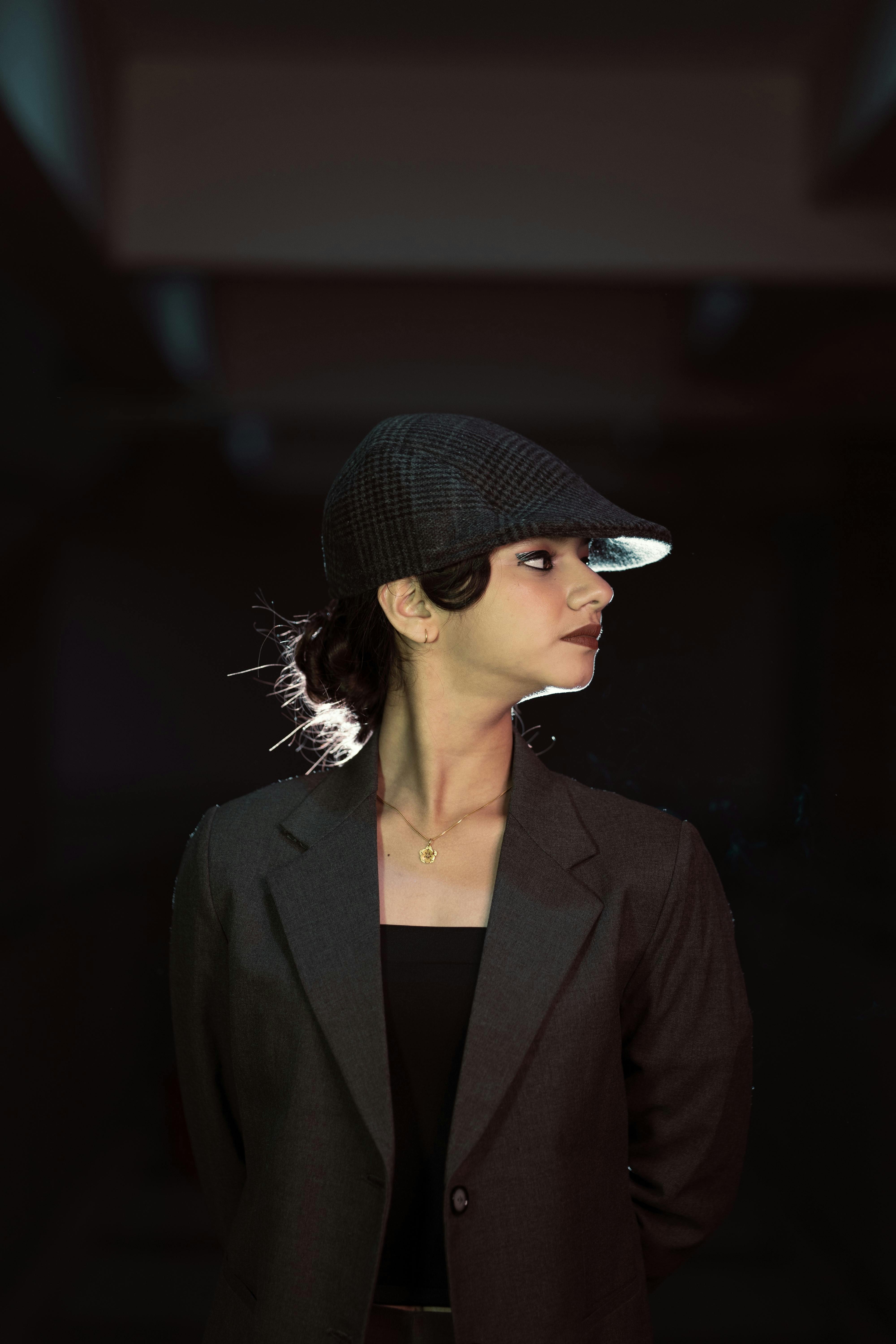 Gratis Elegant kvinne i mørk blazer og hatt, ser til siden med dramatisk belysning innendørs. Arkivbilde