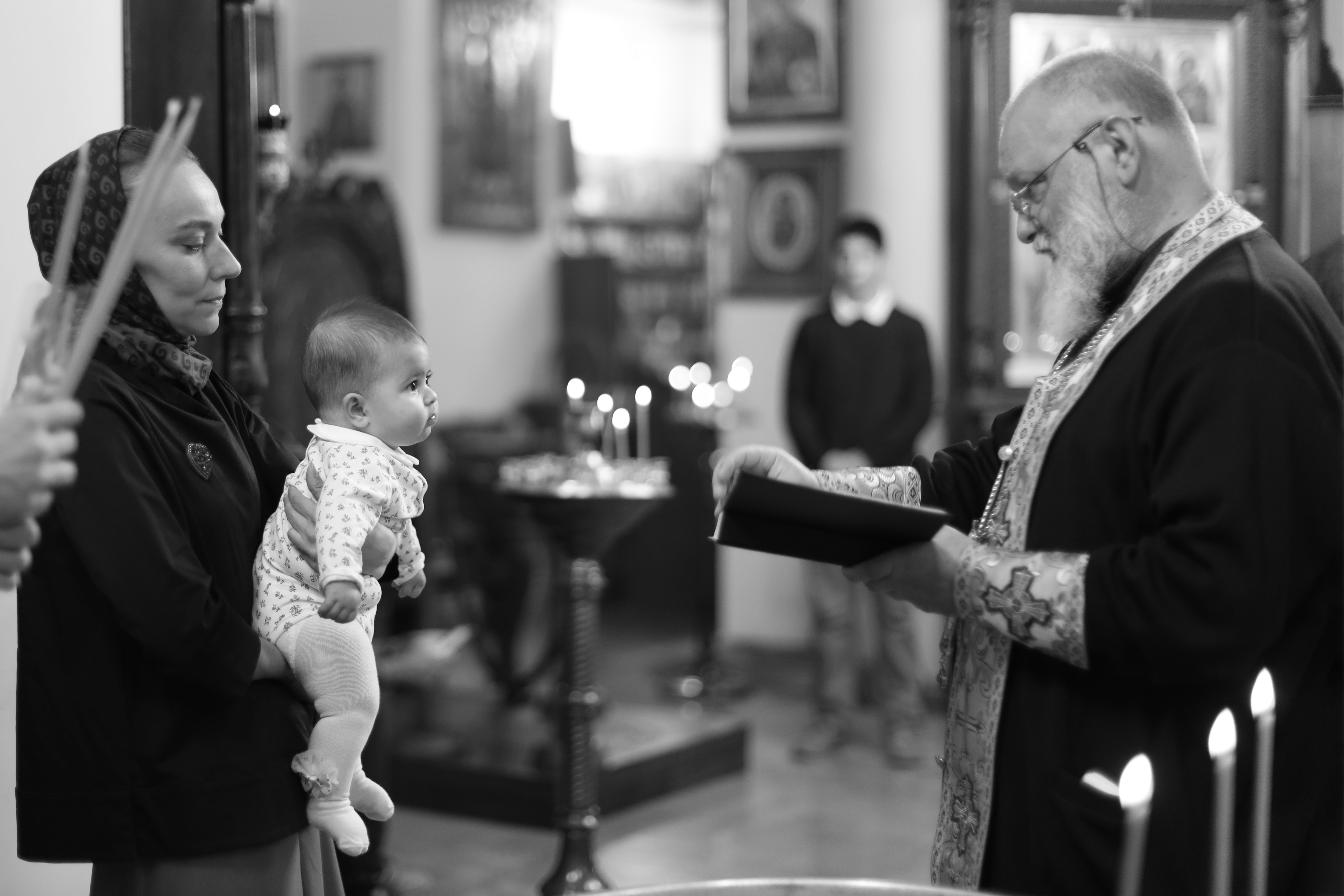 O ceremonie solemnă de botez cu un preot, o mamă și un copil într-un cadru tradițional de biserică.