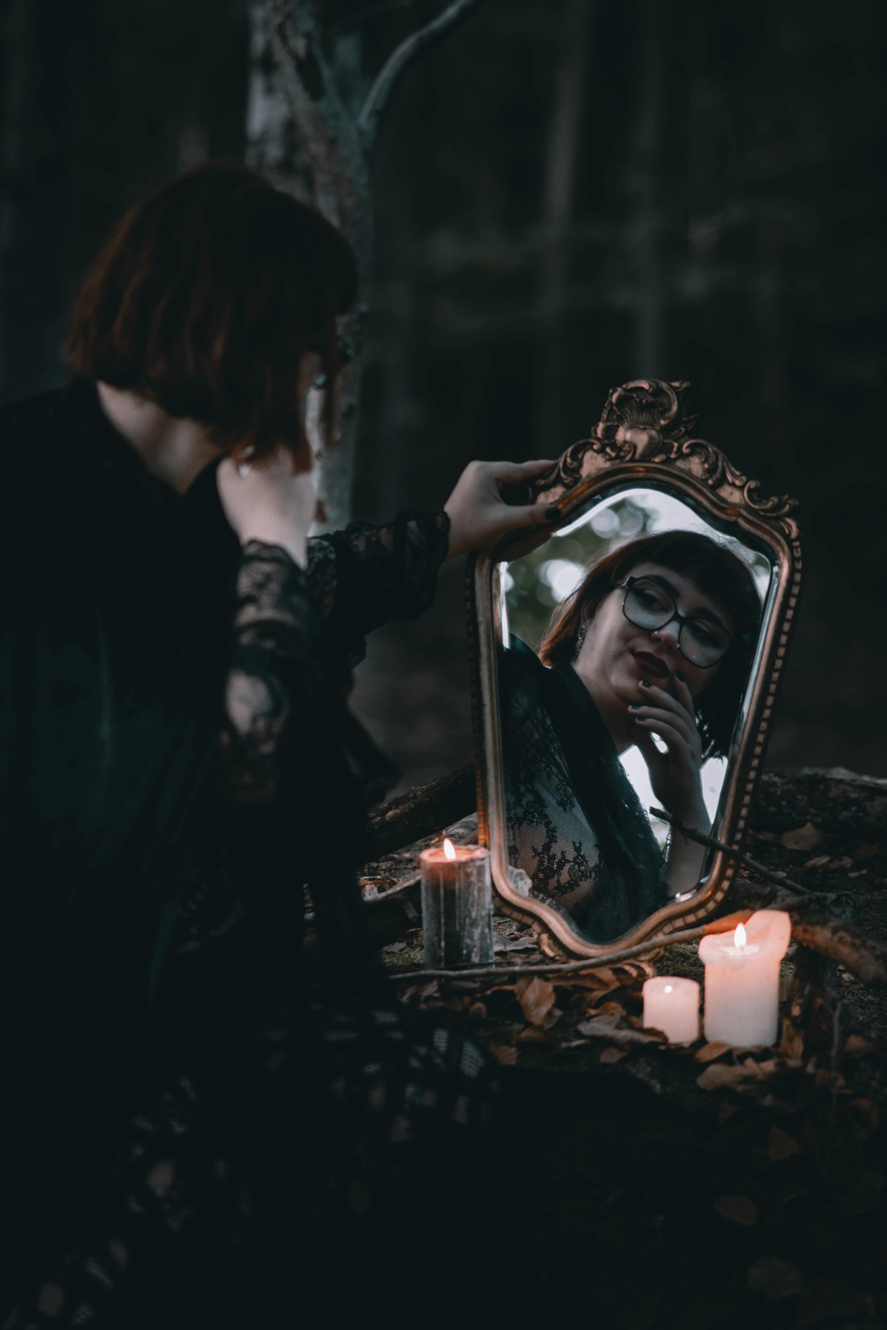A woman gazing into a mirror amidst candles in a dark forest, evoking a magical and mysterious atmosphere.