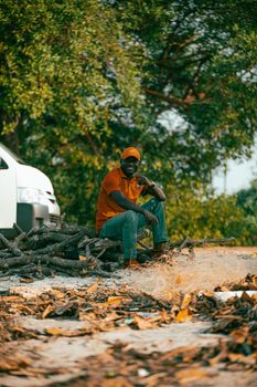 A man in casual attire sits on a pile of logs outdoors, amidst a natural setting.