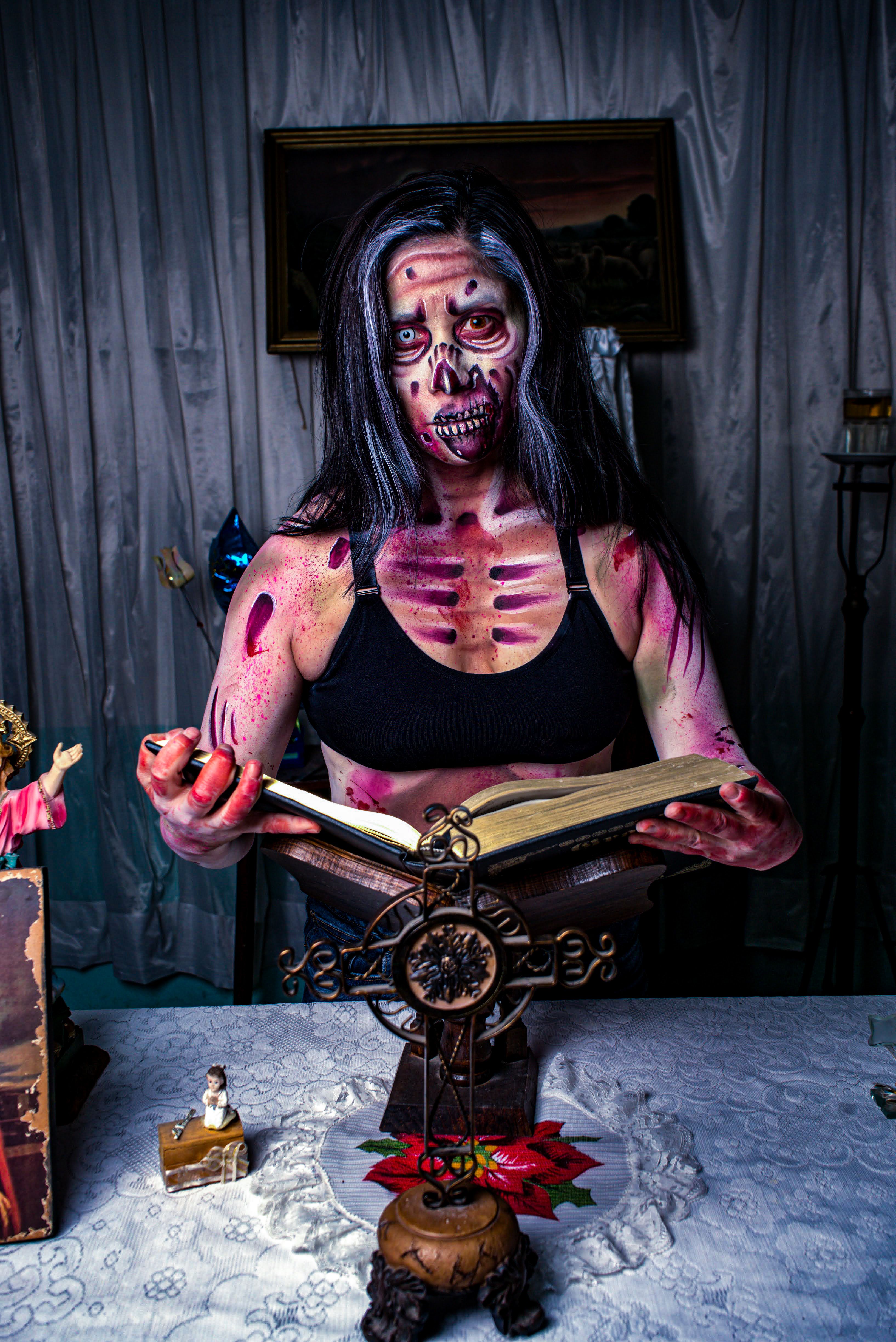 A woman with terrifying skeletal makeup reads an ancient book in a chilling, dimly lit room.